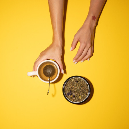 Main tenant une tasse avec une boule à infusion et un bocal de fleurs de CBD à côté